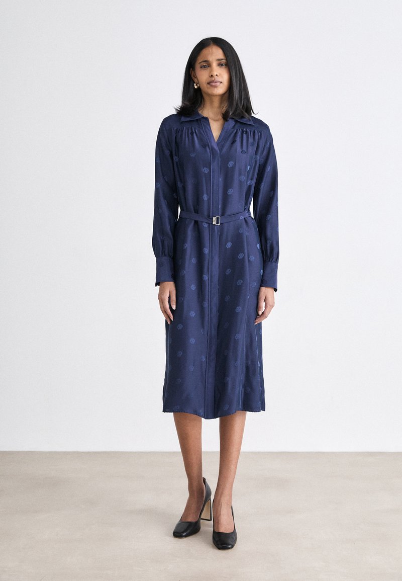 Abito camicia blu navy con maniche lunghe, motivo testurizzato, vita stretta con una cintura, lunghezza al ginocchio, abbinato a tacchi neri.