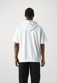 Bijela kratka majica s kapuljačom i vezicom, s opuštenim krojem i elastičnim rubom. Nosena s crnim hlačama, prikazuje stražnji pogled.