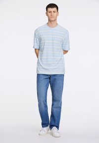 Ljusblå randig t-shirt med korta ärmar, kombinerad med blå denimjeans och vita sneakers, stående mot en enfärgad vit bakgrund.