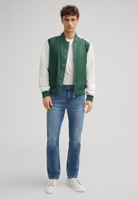 Giacca bomber verde con maniche bianche, abbinata a jeans blu strappati e sneakers bianche. La giacca presenta una chiusura con zip e polsini a coste.