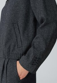 Dunkelgrau maßgeschneiderte Jacke aus strukturiertem Material, mit einer Seitenasche und einem knöpfbaren Ärmel. Elastischer Bund für eine körpernahe Passform.