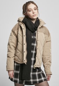 Beige gesteppte Jacke mit Fellakzenten an den Schultern, kombiniert mit einem schwarzen Schal über einem schwarz-weißen Karokleid.