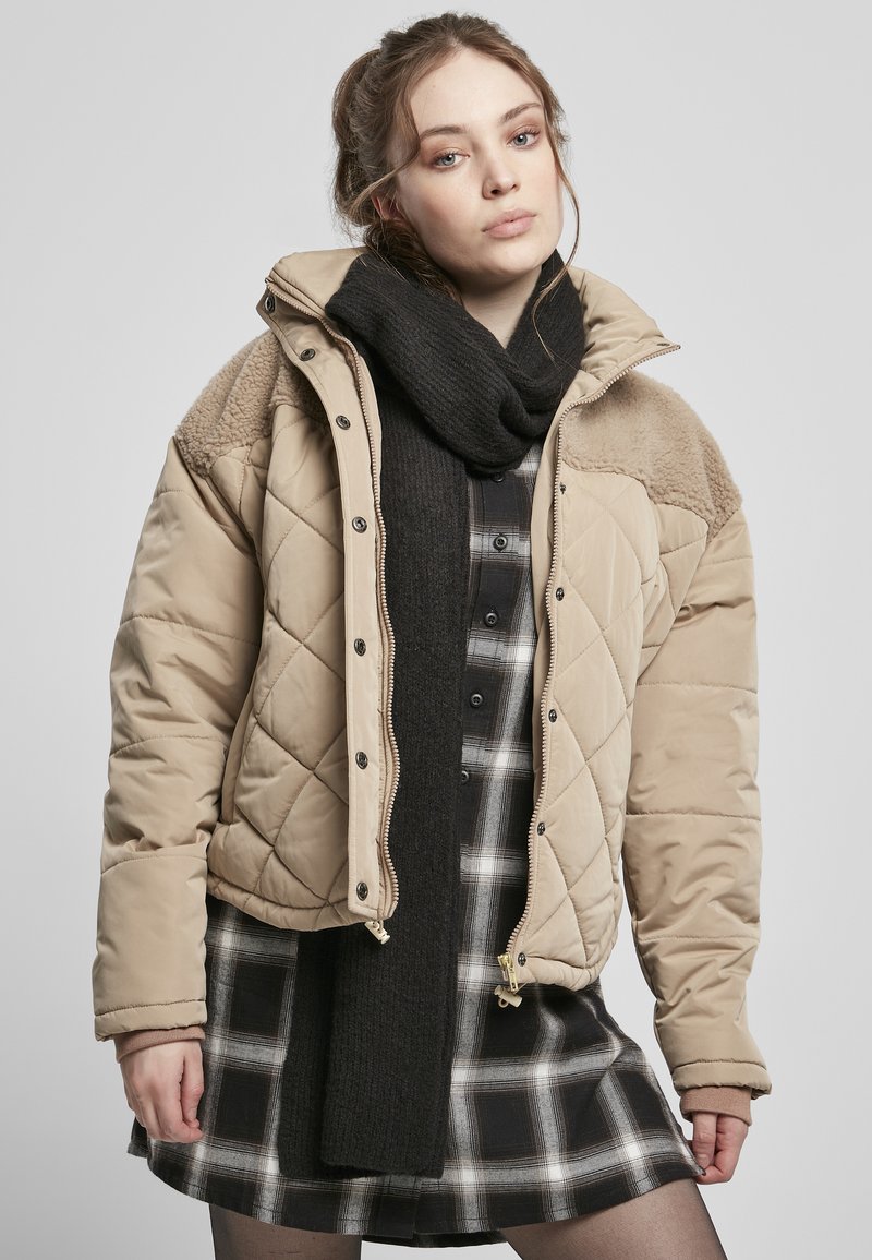 Beige gesteppte Jacke mit Fellakzenten an den Schultern, kombiniert mit einem schwarzen Schal über einem schwarz-weißen Karokleid.
