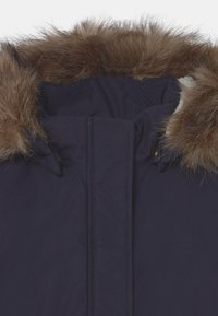 Parka bleu marine avec un col en fausse fourrure marron amovible, présentant une texture matelassée et une coque extérieure lisse. Accents dorés.