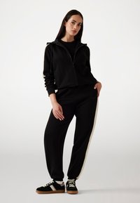 Crna zip-up hoodie s bočnim džepovima, uparena s crnim trenirkama s kremastom prugom. Model nosi crno-kremaste tenisice.