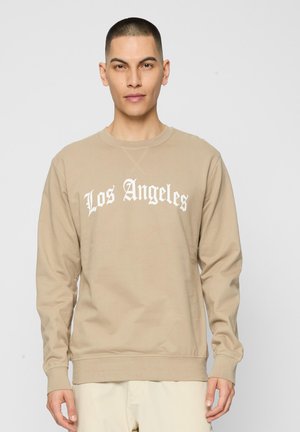 Homme portant un sweat-shirt beige avec l'inscription "Los Angeles" en police gothique blanche, debout devant un fond gris clair uni.