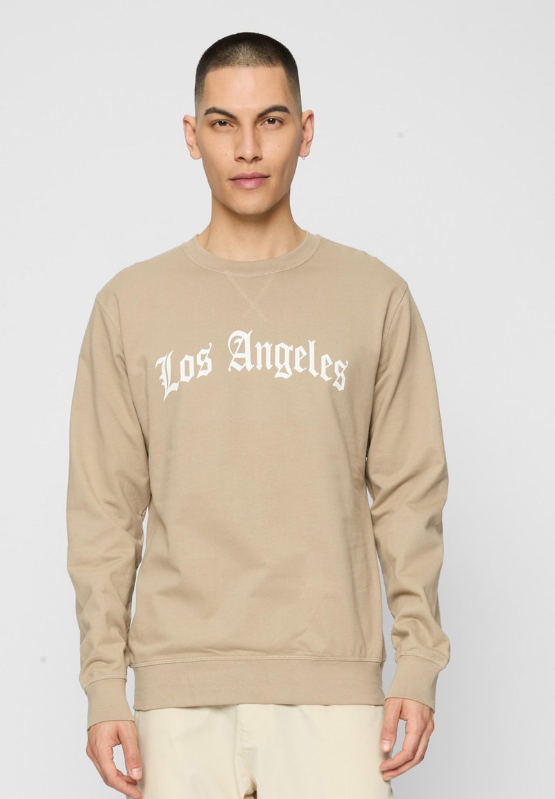 Άνδρας με μπεζ φούτερ που γράφει «Los Angeles» με λευκά γοτθικά γράμματα, στέκεται μπροστά σε ένα μονόχρωμο ανοιχτό γκρι φόντο.