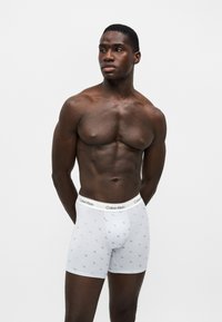 Homme musclé à la peau foncée, debout, les bras dans le dos, portant un boxer blanc Calvin Klein avec un motif logo sur fond uni.