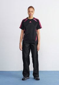 Μαύρο αθλητικό μπλουζάκι με ροζ λεπτομέρειες και κοντά μανίκια, σε συνδυασμό με γυαλιστερούς μαύρους χαλαρούς παντελόνες και μαύρα αθλητικά παπούτσια.