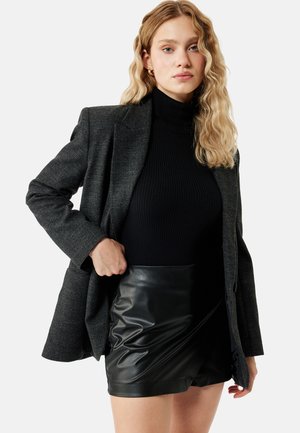 Młoda kobieta z falującymi blond włosami, ubrana w ciemny kraciasty blezer, czarny prążkowany golf oraz czarną skórzaną spódnicę na jednolitym tle.