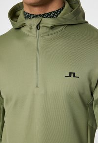 Oliivivihreä vetoketjuhuppari, jossa on korkea kaulus, valmistettu sileästä ja venyvästä kankaasta; siinä on musta logo rinnassa ja sauman yksityiskohtia.