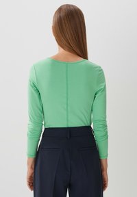 Langsleeve, aansluitend top in lichtgroene stof, met een ronde halslijn en subtiele achterkantnaad. Gepaard met donkere broeken.