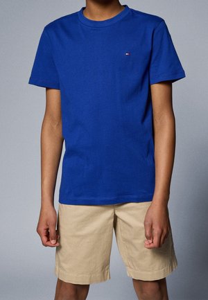 Personne portant un t-shirt bleu à manches courtes avec un petit logo sur la poitrine et un short beige jusqu'aux genoux sur un fond gris uni.