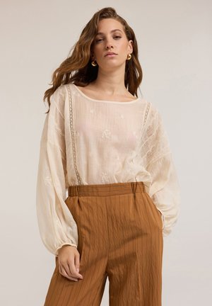 Lichtbeige blouse met geborduurde bloemenpatronen, brede mouwen en kantdetails, gecombineerd met bruine plooibroek.