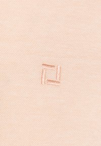 Helle Pfirsich-Strickware mit einem kleinen, gestickten quadratischen Logo in einem etwas dunkleren Farbton. Die Textur ist glatt und gleichmäßig.