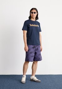 Jūras spēku kokvilnas t-krekls ar dzeltenu "Timberland" logotipu, kombinēts ar purpura krāsas tīrīšanas šortiem un pelēkdzeltenām sporta apaviem, papildināts ar saulesbrillēm.