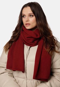 Sciarpa a maglia bordeaux, texture morbida, lunga e drappeggiata attorno al collo, abbinata a un giubbotto imbottito beige chiaro con dettagli sui bottoni.