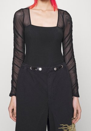 Personne portant un haut noir à encolure carrée avec des manches longues transparentes et un pantalon noir taille haute avec un motif doré près de l'ourlet.