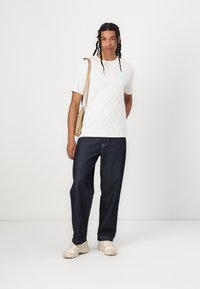 Hvid kortærmet T-shirt med et mintgrønt logo, parret med mørkeblå brede jeans og beige sneakers, bærende en tan crossbody taske.