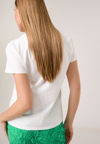 Weißes Kurzarmoberteil, lockere Passform, glatte Textur, getragen mit einem grünen Blumenspitzenrock, von hinten sichtbar. Lange, gerade Haare.