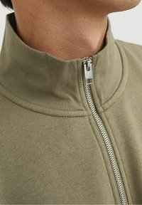 Olivgrön zip-jacka med hög krage och silverdetalj, i en strukturerad bomullstextil med kontrasterande sömmar.