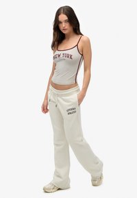 Grijze tanktop met "NEW YORK" print en bordeauxrode rand, gecombineerd met witte joggingbroek met "SUPERDRY ATHLETICS" in zwarte letters.