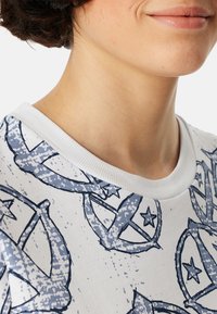 Weißes T-Shirt mit blauem Grafikdesign, das Sterne und abstrakte Formen zeigt. Gerippter Rundhalsausschnitt; glatte Baumwolltextur.