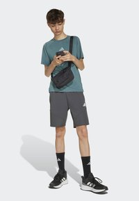 Tyrkysové športové tričko, šedé šortky s akcentmi, čierna crossbody taška, čierne tenisky a ponožky. Model drží telefón a stojí na jednoduchom pozadí.