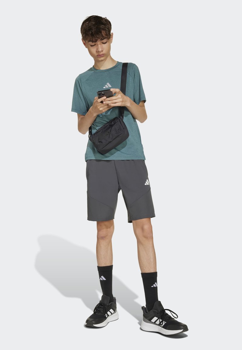 Tyrkysové športové tričko, šedé šortky s akcentmi, čierna crossbody taška, čierne tenisky a ponožky. Model drží telefón a stojí na jednoduchom pozadí.