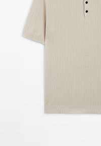 Polo en maille beige à manches courtes avec texture côtelée, trois boutons noirs et un ourlet côtelé sur fond blanc.