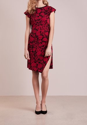 Kobieta w czerwonej kwiecistej sukience z rozcięciem do uda i czarnych szpilkach, stojąca na tle gładkiego beżowego tła.