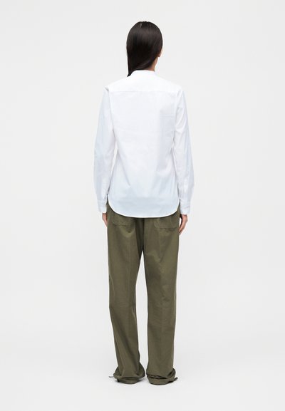 Λευκό πουκάμισο με κουμπιά, μακρυά μανίκια και στρογγυλεμένη γραμμή στο κάτω μέρος, σε συνδυασμό με φαρδιά χακί παντελόνια. Απλός σχεδιασμός χωρίς ορατά σχέδια.