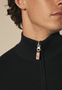 Crni zip-sveta sa visokim ovratnikom, koji ima teksturirani uzorak pletenja i srebrni zip koji je ukrašen tamno smeškim kožnim povlačnikom.