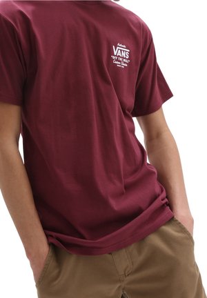 Persoon draagt een bordeauxrode Vans "Off The Wall" T-shirt met handen in de zakken van bruine broek, zichtbaar vanaf de schouders tot de heupen.