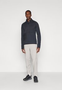 Pullover zippé navy en tissu texturé avec une poche noire ; associé à un pantalon cargo beige clair et des chaussures de sport noires.