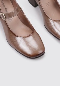 Zapatos de cuero marrón con tacón cuadrado y puntera cuadrada, con una correa estrecha en el tobillo y una superficie texturizada. Sin patrones o acentos visibles.