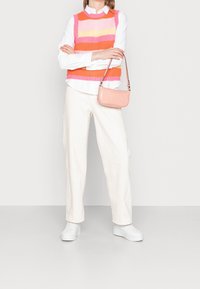 Person står med armene korslagte iført en farverig stribet ærmeløs sweater, hvid skjorte, hvide bukser, hvide sneakers og holder en lyserød håndtaske.