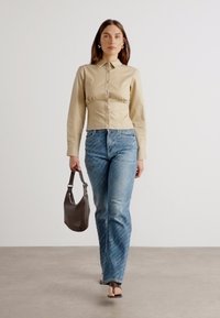 Camisa ajustada beige con mangas largas y cintura fruncida, combinada con jeans de mezclilla azules con costuras blancas. Los accesorios incluyen un bolso oscuro.