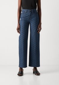 Blaue weite Jeans mit wiederholtem DKNY-Logo-Muster, hohem Bund und klassischem Knopfverschluss, kombiniert mit schwarzen Blockabsatzsandalen.