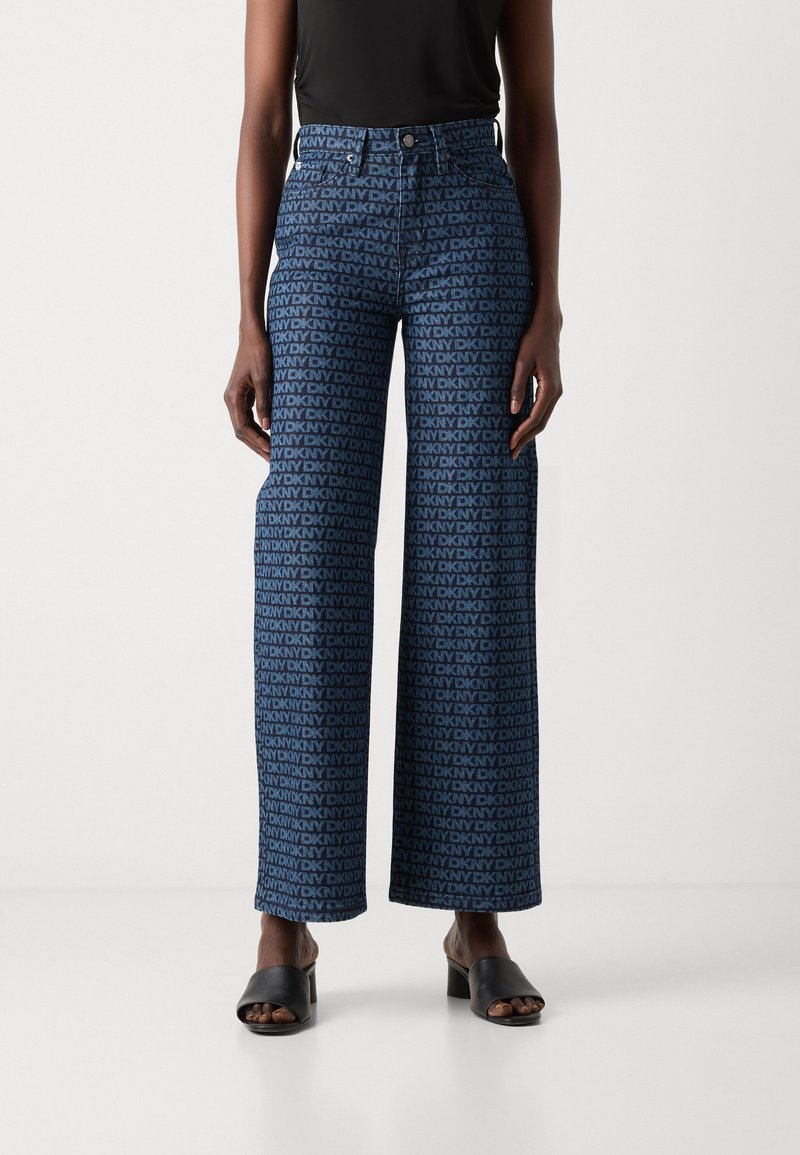 Jeans de pierna ancha azules con un patrón repetido del logo de DKNY, con cintura alta y cierre clásico de botón, combinados con sandalias negras de tacón cuadrado.