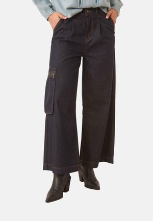 Persona con pantalones anchos de mezclilla oscura con un bolsillo lateral con cremallera, suéter gris claro y botines negros de tacón.