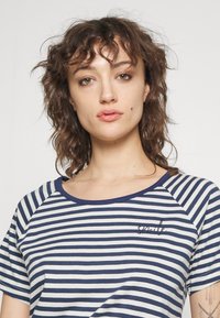Camiseta de rayas en azul marino y blanco, con cuello redondo y mangas cortas, que presenta un detalle bordado de "sonrisa" en el pecho. Textura de tejido suave.
