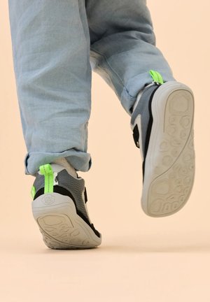 Kind draagt grijze en zwarte sneakers met neon groene treklabels, loopt met één voet opgetild, lichtblauwe broek opgerold bij de enkels.