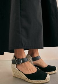 Zoccoli espadrillas neri con suola in juta, punta chiusa e retro intrecciato. Presentano un cinturino alla caviglia nero con una fibbia in argento.