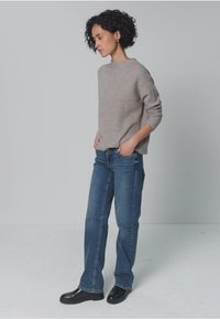 Suéter de punto gris claro con mangas remangadas, combinado con jeans de mezclilla azul. El modelo está de perfil, con las manos en los bolsillos y lleva zapatos negros.