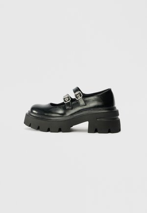 Pantofi cu platformă din piele neagră, cu vârful rotunjit, două accesorii din cataramă metalică și o talpă chunky, texturată, tot neagră.
