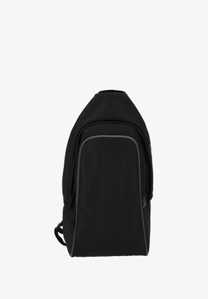 Lyshåret kvinde med hestehale iført sort outfit og hvide sneakers, bærende på en sort rygsæk, vendt væk på en ensfarvet hvid baggrund.