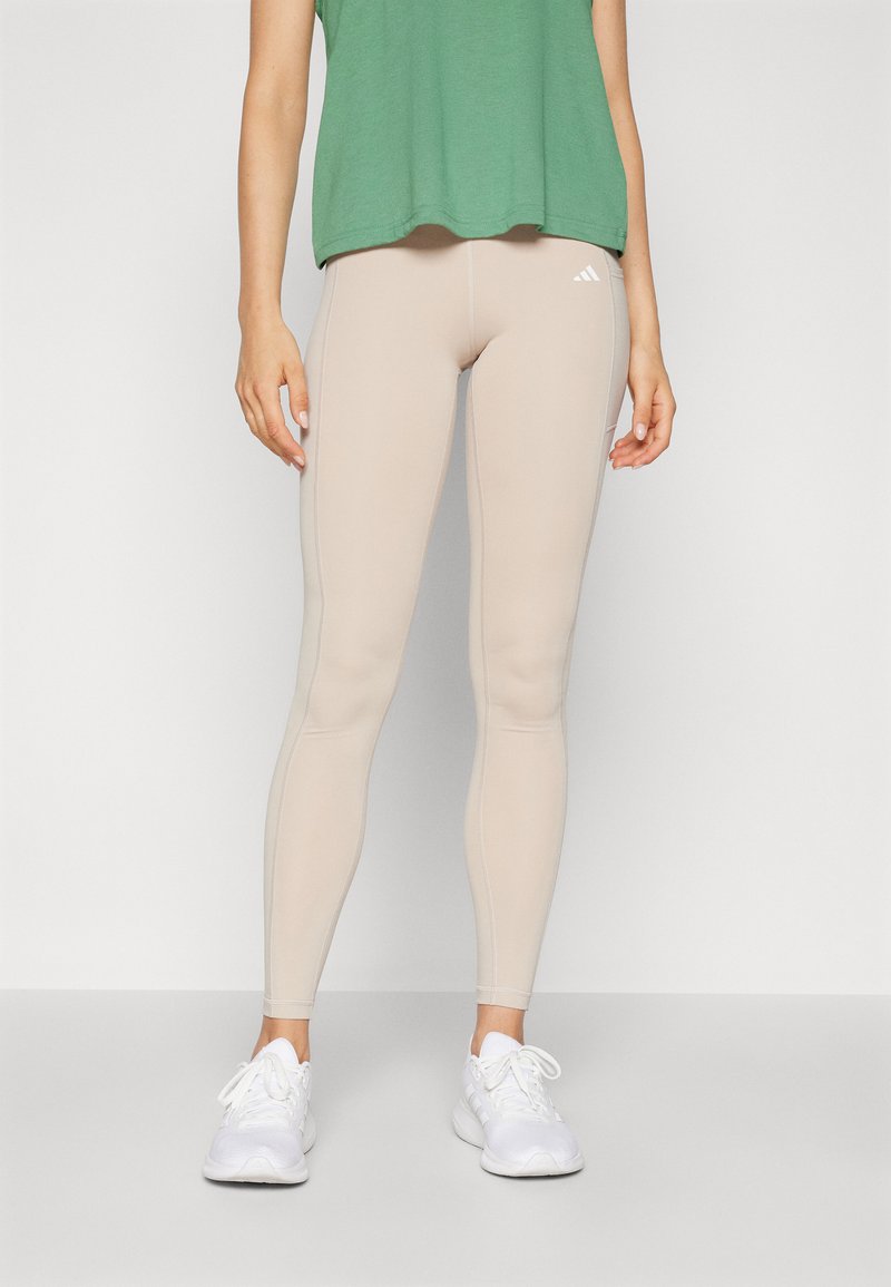Leggings beige con una textura suave y elástica, con acentos laterales en blanco y un logo de Adidas. Combinados con una camiseta verde holgada y zapatos blancos.