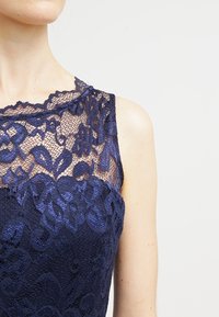 Femme portant une robe sans manches en dentelle bleu marine avec des motifs floraux et un col transparent sur un fond clair uni.