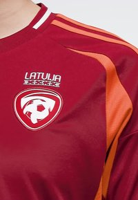 Rode en oranje korte mouwen trui met de tekst "LATVIJA" boven een voetbalembleem; heeft gestreepte schouderaccenten en een gestructureerde stof.